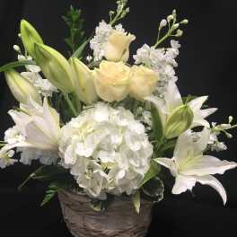 White lilies and pale roses in a woven basket with a large white hydrangea bloom