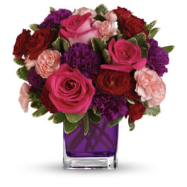 Compact bouquet of hot pink roses, red and purple flowers in a purple glass cube vase