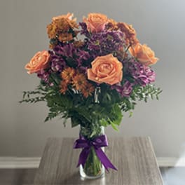 Peach roses and purple flowers in a clear glass vase with a purple ribbon