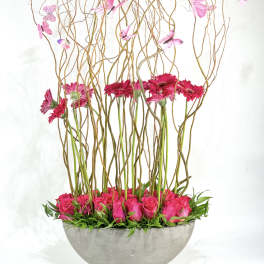 Pink roses and gerbera daisies in a gray bowl with tall curly branches and butterflies