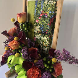 Floral arrangement with orange roses, purple lilacs, and dark calla lilies on an easel