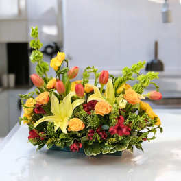Mixed floral arrangement with yellow lilies, orange roses, and red tulips in a low container