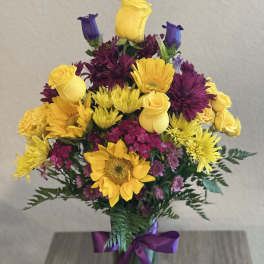 Bouquet of yellow roses, sunflowers, and purple blooms in a glass vase