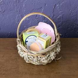 Woven gift basket filled with boxed soaps and a pink scrub brush on a wooden table