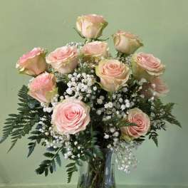 Tall arrangement of light pink roses and white filler flowers in a clear glass vase on a small dark table.