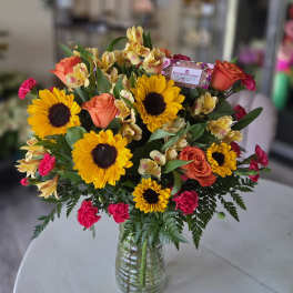 Vase arrangement of bright yellow sunflowers, orange roses, pink carnations, and yellow alstroemeria.