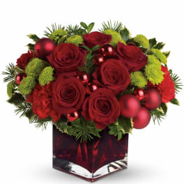 Low red and green holiday arrangement with red roses, carnations, ornaments, and evergreens in a red cube vase