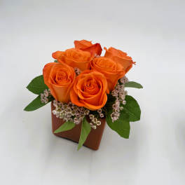 Orange roses arranged in a small brown square vase with pink filler flowers