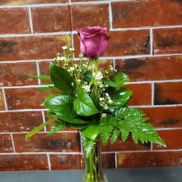 Single purple rose in a clear glass vase with greenery and small white filler flowers