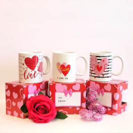 Three Valentine mugs on heart-patterned gift boxes with a red rose and glitter heart.