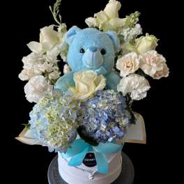 Blue teddy bear surrounded by white roses and blue hydrangeas in a gift box