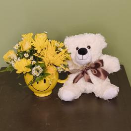 Yellow flower arrangement in a smiley-face mug beside a white teddy bear
