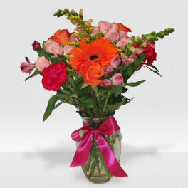Mixed bouquet of bright orange, pink, and red flowers in a clear vase with a pink ribbon
