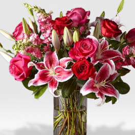 Tall arrangement of pink lilies and red roses in a clear vase with a gold band