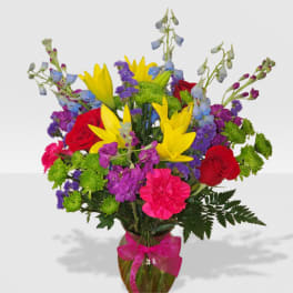 Bright mixed bouquet of yellow lilies, red roses, pink carnations, and purple blooms in a glass vase with pink bow