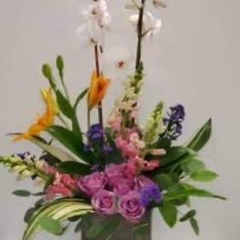 Tall mixed floral arrangement in a dark square vase with pink, purple, white, and orange blooms