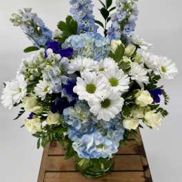 Blue hydrangeas, white daisies, and cream roses arranged in a clear glass vase.