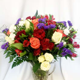 Mixed bouquet of roses, carnations, and purple filler flowers in a glass pitcher vase