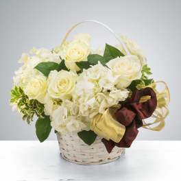 White roses and hydrangeas in a white wicker basket with a gold and burgundy ribbon bow