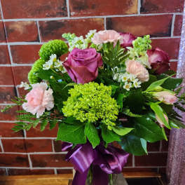 Mixed bouquet of roses, carnations, and green blooms in a glass vase with a purple ribbon