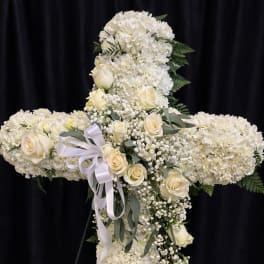 Tall white floral cross on an easel with roses and a white ribbon bow.