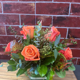 Orange roses arranged in a square glass vase with mixed greenery