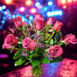 Arrangement of pink roses with small purple blooms in a green glass vase on a reflective surface.