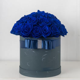 Round box arrangement of vivid blue roses in a dark hatbox with a blue ribbon and rhinestone accent.