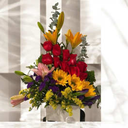Mixed bouquet with red roses, yellow lilies, and sunflowers in a white container