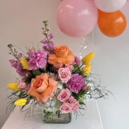 Pink and orange flower arrangement with balloons in a glass vase