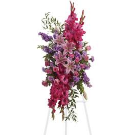 Standing spray of hot pink gladiolus, pink lilies, and lavender flowers on a white easel.