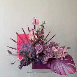 Purple floral arrangement with roses, orchids, and tropical leaves in a pink container