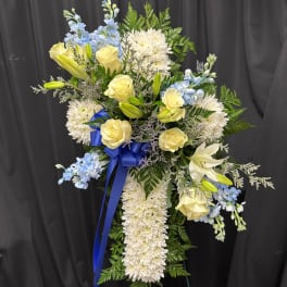Standing spray of white mums, yellow roses, lilies, and blue flowers with blue ribbon on an easel.
