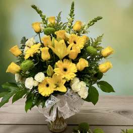 Yellow roses, lilies, and gerbera daisies in a glass vase with a white ribbon.