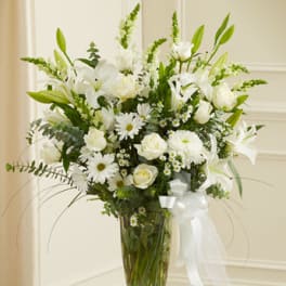 Tall arrangement of white lilies, roses, daisies, and mums in a clear glass vase with a white bow