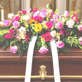 Bright pink and cream rose casket spray with white ribbon on a wooden casket.