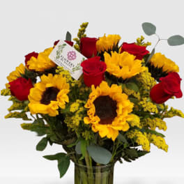 Tall vase arrangement of yellow sunflowers and red roses with yellow filler flowers and a small card