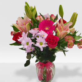 Mixed bouquet of pink lilies, red roses, and other blooms in a clear glass vase with a pink bow.