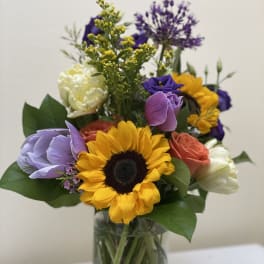 Mixed bouquet with a sunflower, roses, and purple blooms in a glass vase