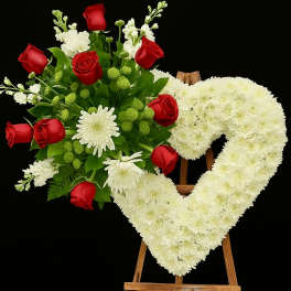 Heart-shaped white floral wreath on an easel beside a red rose bouquet