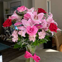 Pink lilies and roses arranged in a clear glass vase with a pink ribbon.