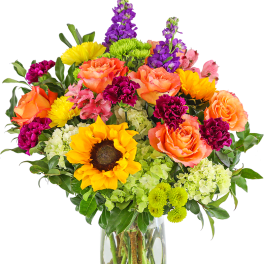 Tall mixed bouquet with sunflowers, orange roses, green hydrangeas and purple stock in a clear vase.