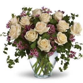 Ivory roses with small purple blooms and trailing ivy in a clear glass vase.
