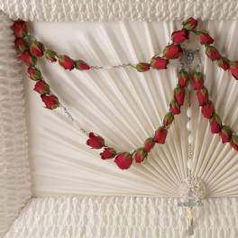 Red rosebud rosary draped across an ivory pleated casket interior with crucifix