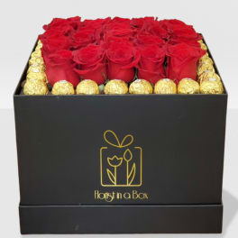 Square black box filled with red roses framed by gold-wrapped chocolates.