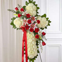 Tall standing cross of white flowers with red roses and lilies on an easel with red ribbon.