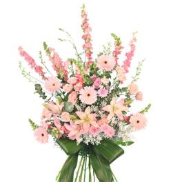 Tall standing spray of pink lilies, gerbera daisies, carnations and snapdragons with large green leaves on an easel