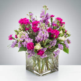 Low arrangement of hot pink and lavender flowers with cream blooms in a clear square glass vase