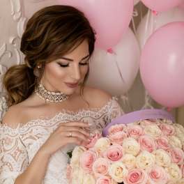 Woman in a white lace dress holding a box of pink and cream roses with balloons behind her