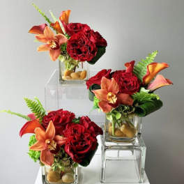 Three small red and orange floral arrangements with orchids and roses in glass cube vases with stones.